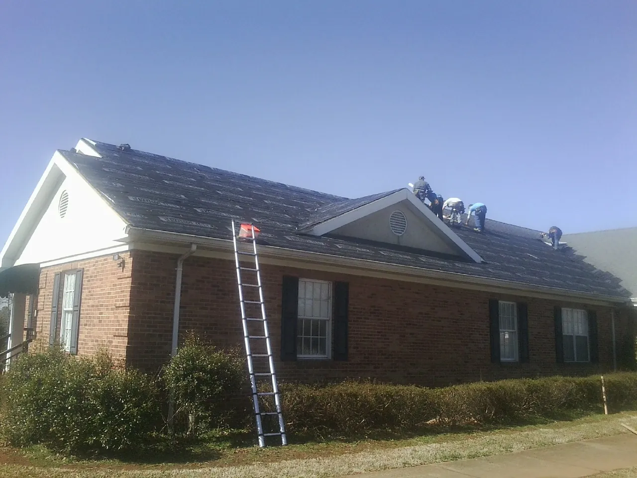 Crew tearing shingles off a brick ranch roof with a ladder set — Emergency Roof Repair work by our crew in Park Valley