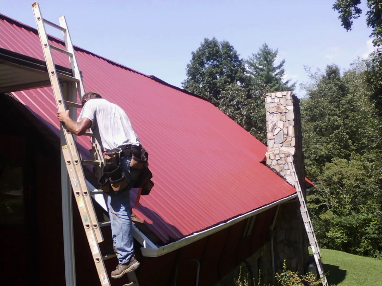 Roofer on a ladder beside a red metal roof and stone chimney — Soffit & Fascia Repair by Roofing Brothers in Park Valley